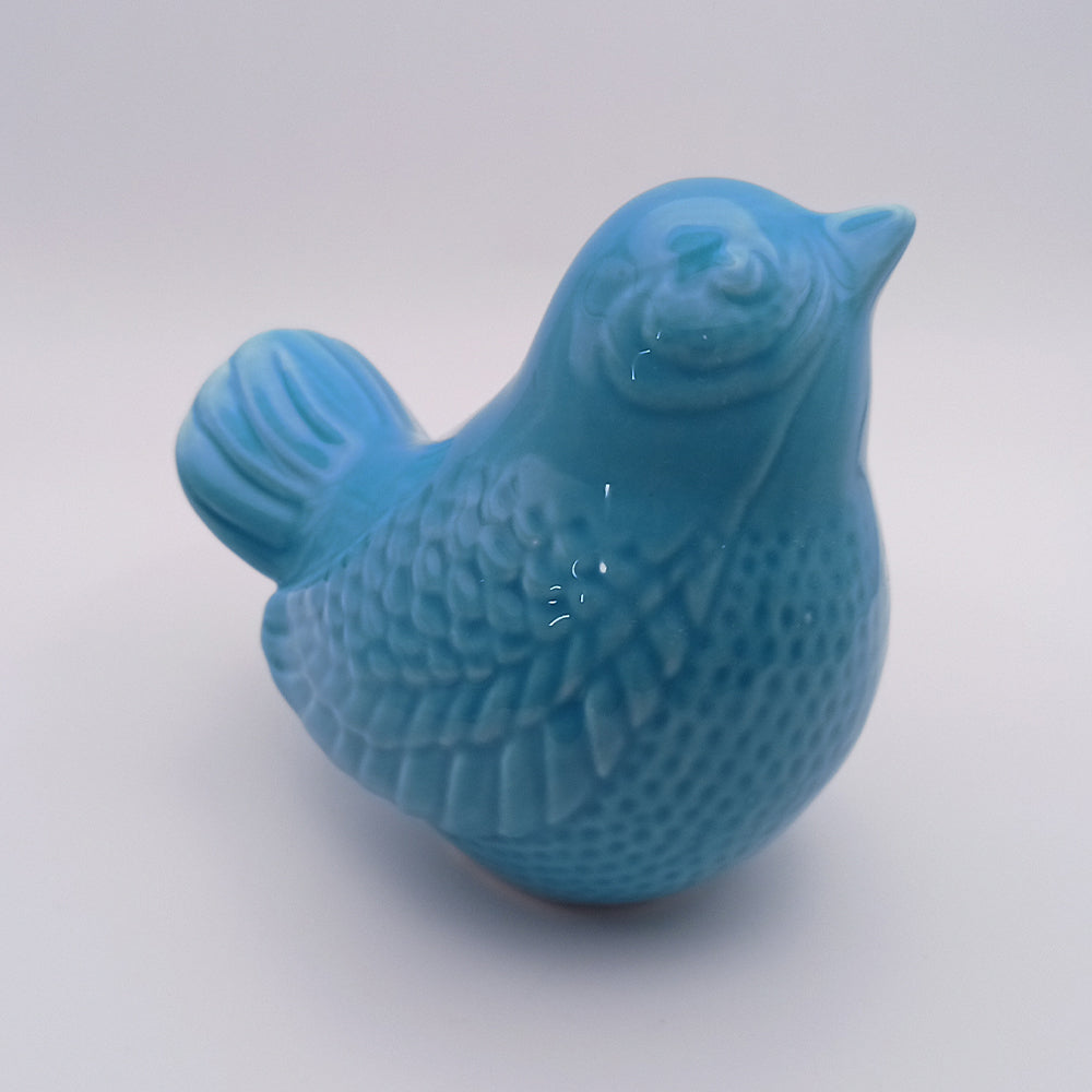 The Tejo Shop's Ceramic Bird Looking Up Figurine is a small, handmade piece colored in glossy light blue. It features detailed textures on the wings and body, and is positioned facing slightly to the right atop a plain white background, making it an exquisite piece of home decor.