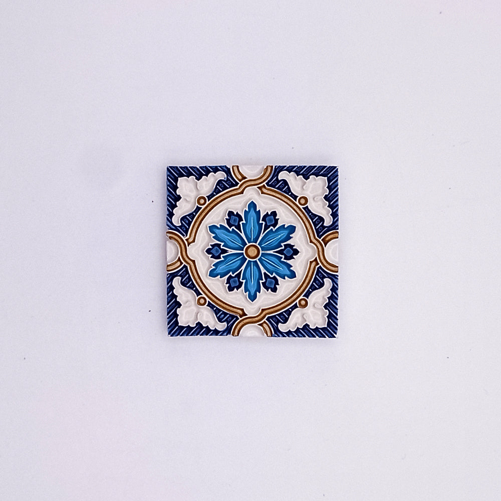 A Tejo Shop small hand-painted ceramic tile with a symmetrical floral design in blue, white, and touches of yellow, centered on a plain light background.