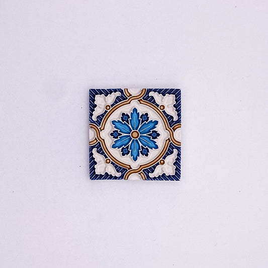 A Tejo Shop small hand-painted ceramic tile with a symmetrical floral design in blue, white, and touches of yellow, centered on a plain light background.