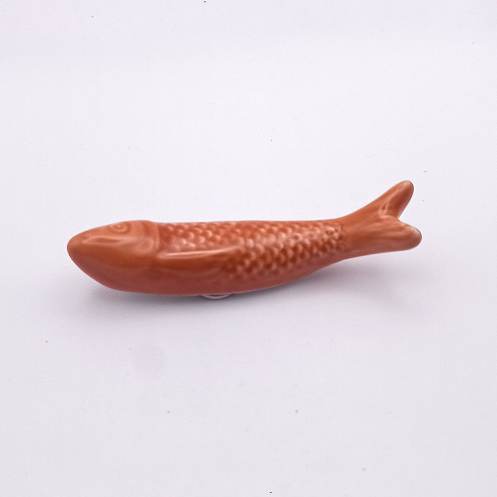 A small Ceramic Sardine Fridge Magnet from Tejo Shop, featuring an orange color with a textured scale pattern and smooth surface, is placed on a white background. This fish figure has a pointed head, streamlined body, and forked tail, resembling a simplified fish shape ideal for kitchen decor.