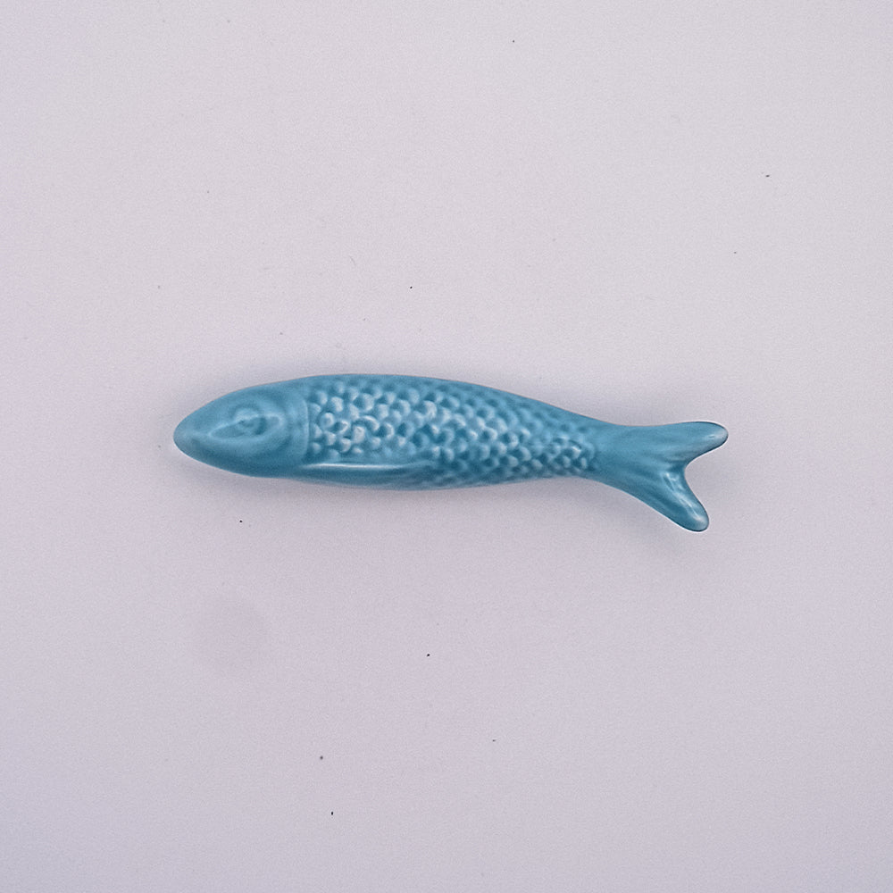 A small blue Ceramic Sardine Fridge Magnet from Tejo Shop, reminiscent of a classic Portuguese icon, is placed against a plain light grey background. The fish features detailed scales and fins, with its head pointing left and tail to the right. This charming ceramic magnet makes an adorable piece of kitchen decor.