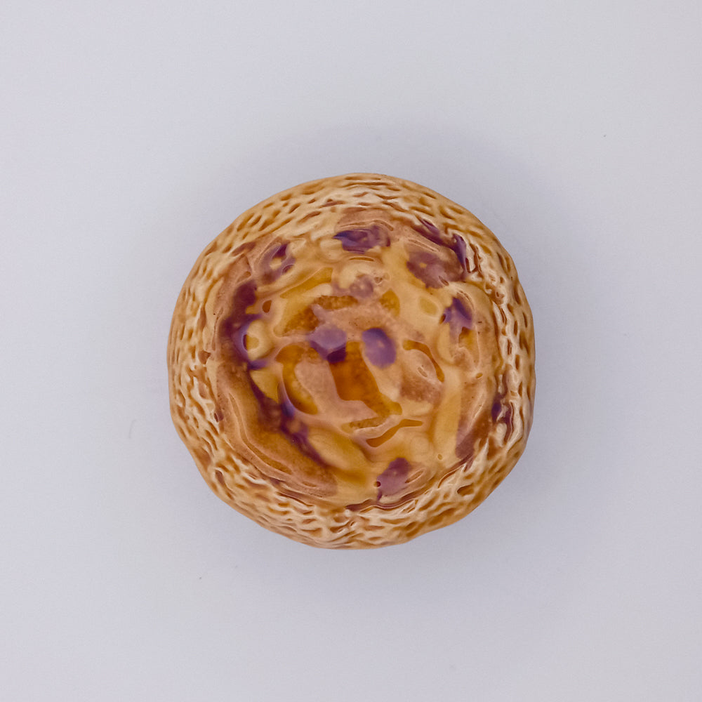 A Ceramic Pastel de Nata by Tejo Shop rests on a plain white background, showcasing a round, golden-brown pastry with a swirled, slightly craggy surface featuring subtle hints of purple. Its textured dimensions create an appealing contrast that draws attention to its delightful form.