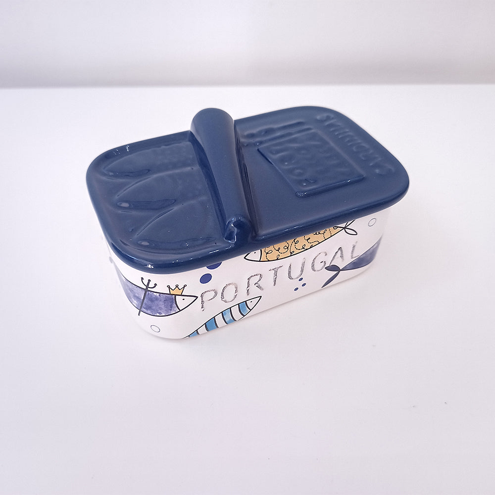 The Ceramic Soap Box - Sardine Special Edition by 100ml is a decorative ceramic container designed to resemble a tin can, featuring ocean-themed illustrations such as fish and waves, with the word "Portugal." This special Portuguese-style edition's navy blue lid complements its white background adorned with vibrant blue and yellow accents.