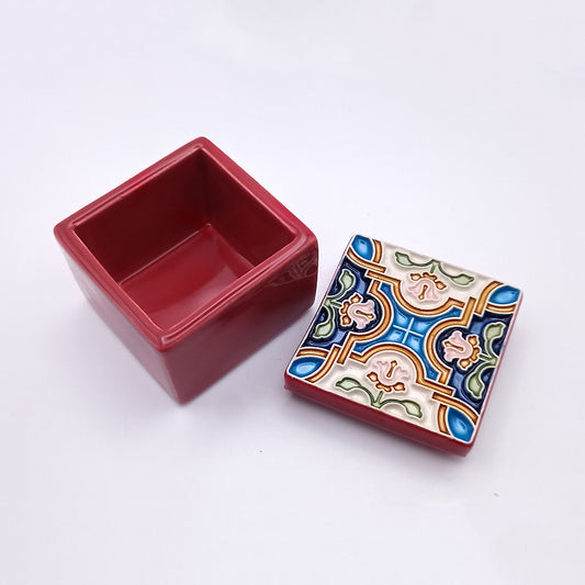 A small, square Coimbra Ceramic Box by Tejo Shop with the lid removed. The box is solid red, while the lid features a colorful, intricate geometric pattern with floral elements in blue, green, and yellow on a white background. This charming ceramic box and its lid are placed on a plain white surface.