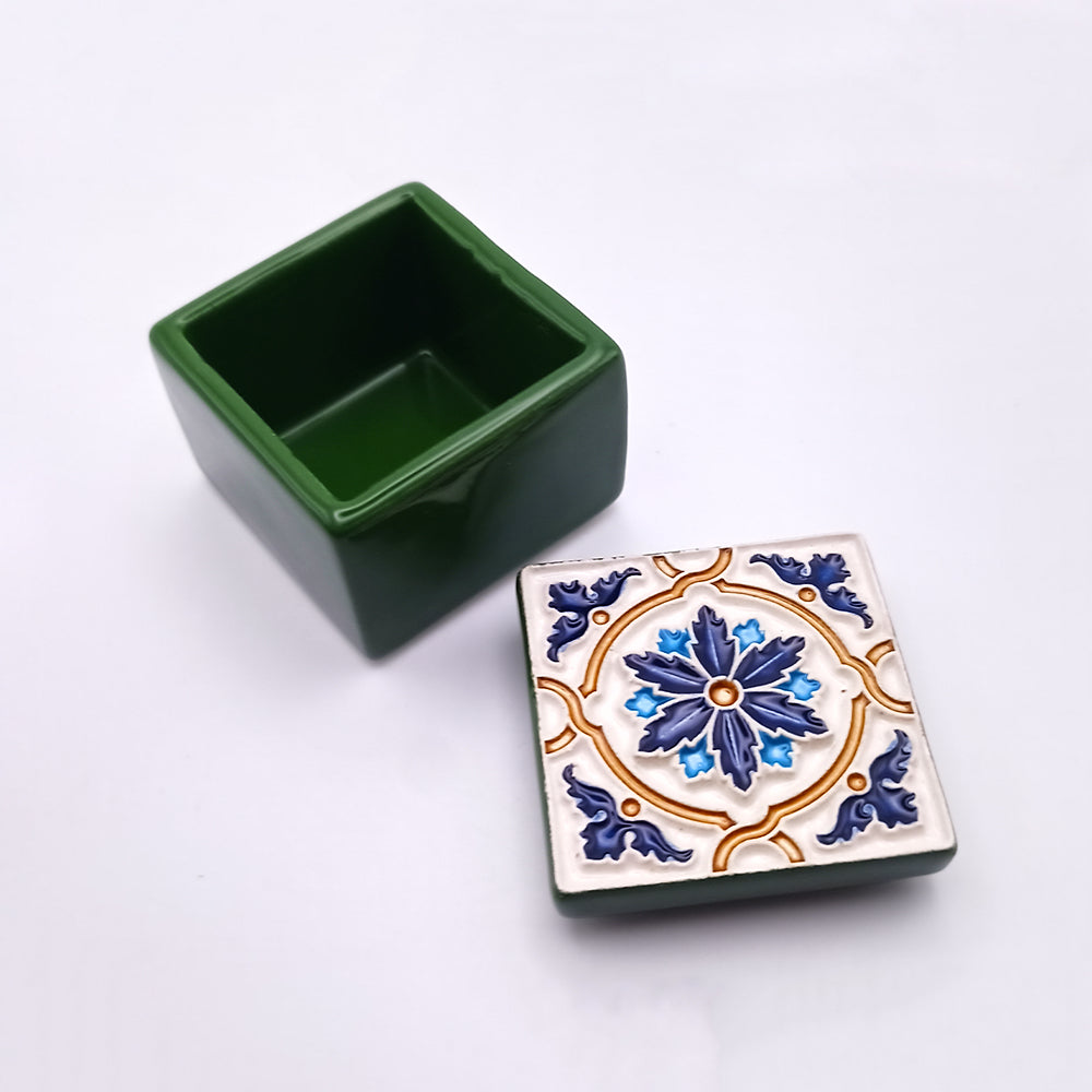 A small, square green container with its lid off. The lid of this Corvo Island Ceramic Box by Tejo Shop features a decorative pattern of a blue and gold flower with blue leaves on a white background. The ceramic masterpiece is empty and sits on a white surface, adding a sophisticated ambiance to any setting.