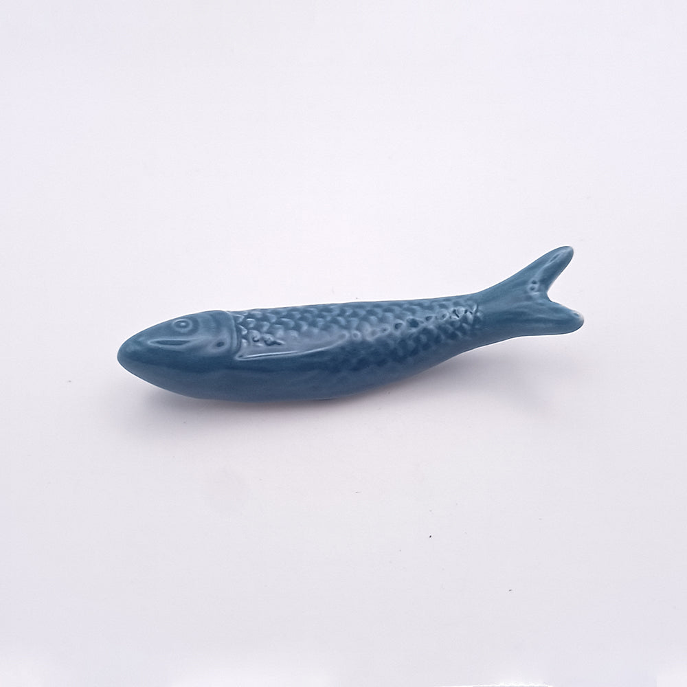 A small, blue ceramic fish with textured scales and smooth fins lies against a plain white background. This charming Ceramic Sardine Fridge Magnet by Tejo Shop serves as both a decorative trinket and an eye-catching piece of kitchen decor, embodying the essence of a beloved Portuguese icon.