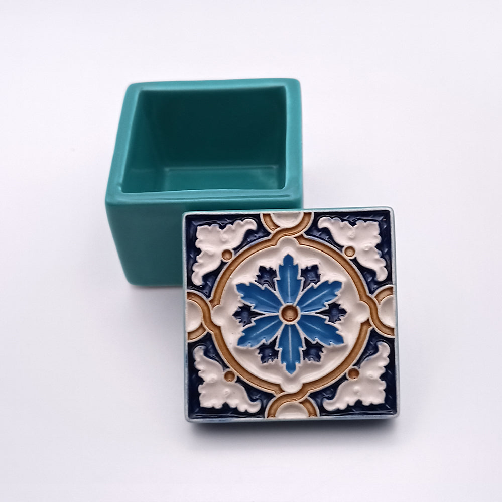 The Discoveries Ceramic Box by Tejo Shop is a square ceramic masterpiece with a removable lid. The box is solid teal with smooth sides, while the lid flaunts an intricate floral and geometric design in blue, white, and tan hues. The box sits open, exuding opulence with its lid leaning elegantly against it.