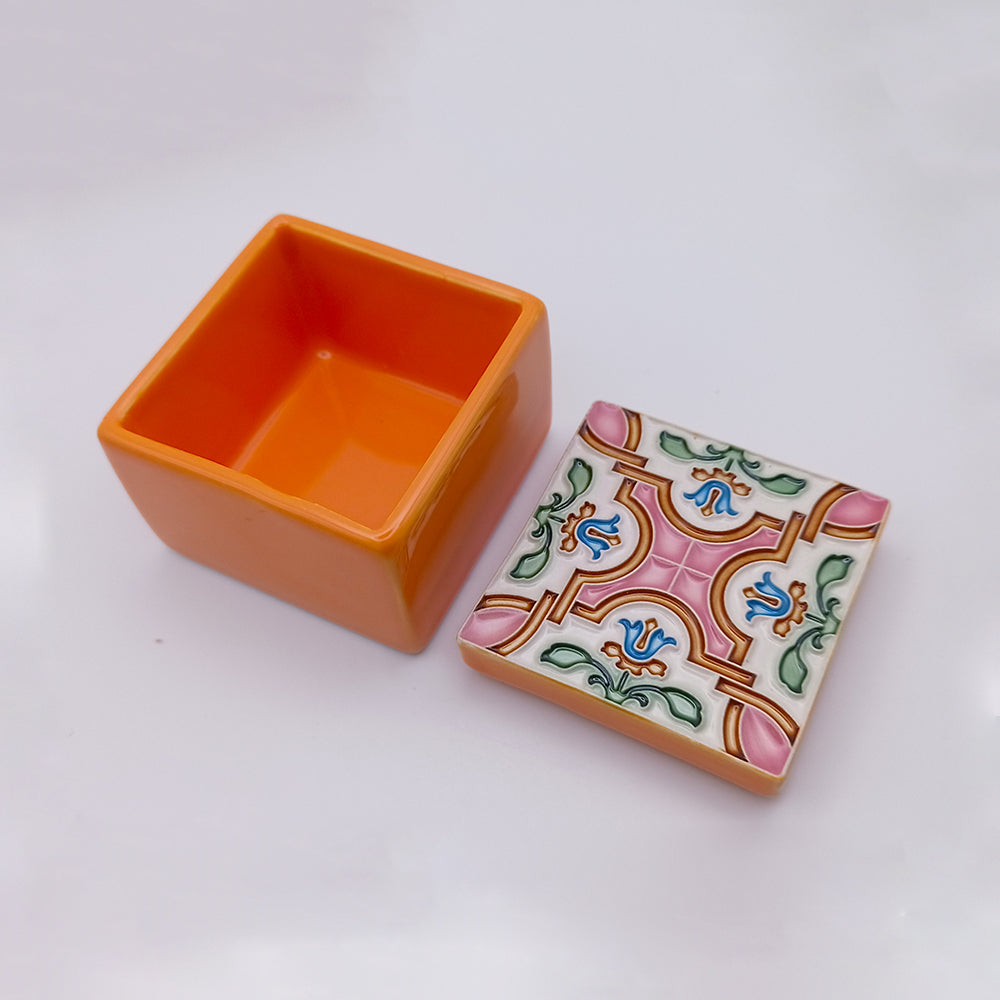 A small, square, orange ceramic box with an intricate, colorful lid design featuring blue floral patterns and pink and green accents. The lid is placed next to the box, revealing its open interior. This Ericeira Ceramic Box by Tejo Shop stands out as a unique ceramic piece against the neutral and unobtrusive background.