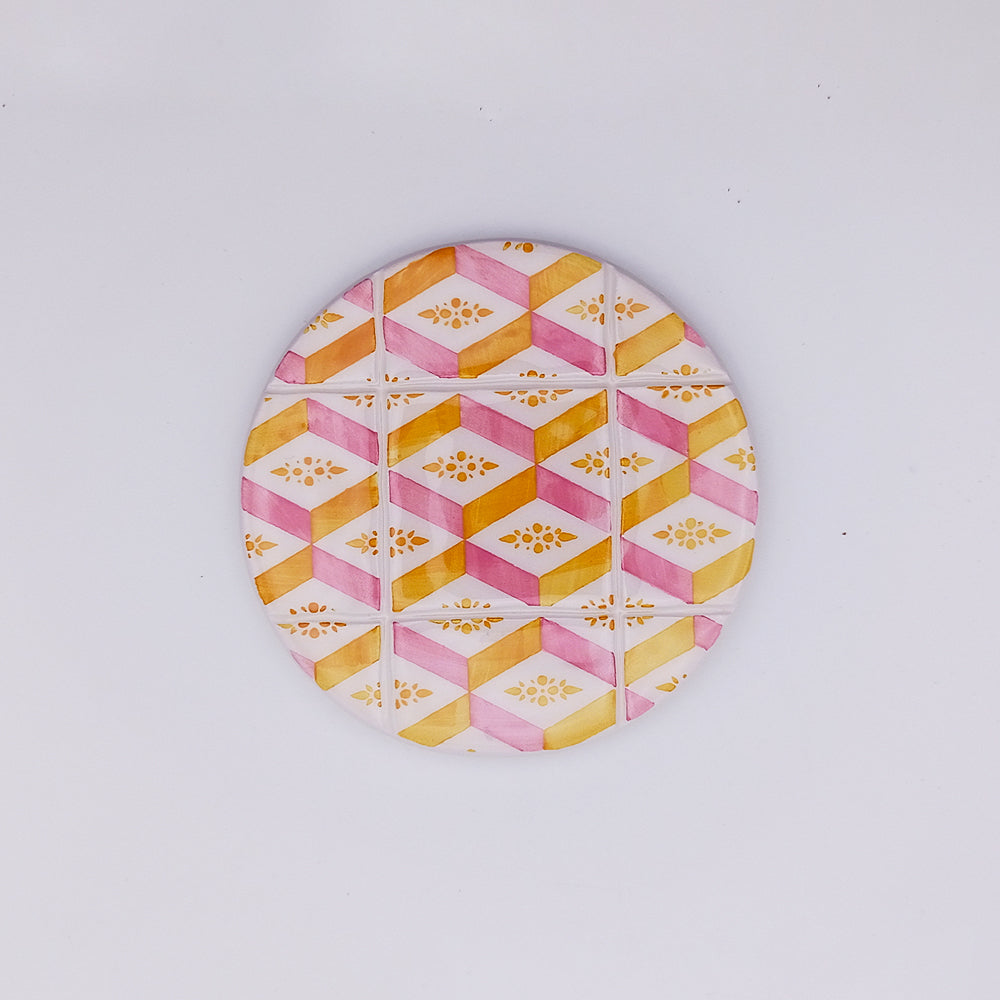 The Tejo Shop Estremoz Ceramic Cup Pad is a circular object with a colorful geometric pattern featuring yellow and pink hexagons arranged in a symmetrical design. Hand-painted ceramic details include intricate floral motifs within the hexagons, all set against a plain white background.