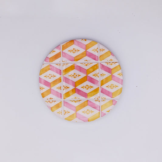 The Tejo Shop Estremoz Ceramic Cup Pad is a circular object with a colorful geometric pattern featuring yellow and pink hexagons arranged in a symmetrical design. Hand-painted ceramic details include intricate floral motifs within the hexagons, all set against a plain white background.