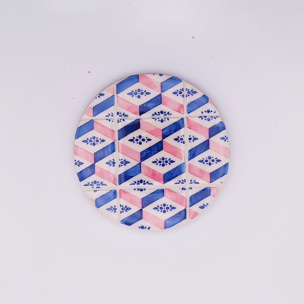 The Tejo Shop Evora Ceramic Cup Pad is a high-quality ceramic tile with a geometric pattern of hexagons in blue and pink hues, featuring floral motifs within the hexagons. The hand-made and hand-painted design creates a three-dimensional cube effect, all set against a plain white background.