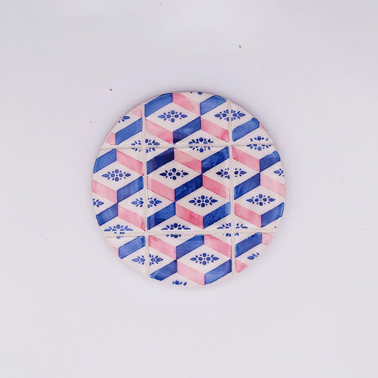 The Tejo Shop Evora Ceramic Cup Pad is a high-quality ceramic tile with a geometric pattern of hexagons in blue and pink hues, featuring floral motifs within the hexagons. The hand-made and hand-painted design creates a three-dimensional cube effect, all set against a plain white background.