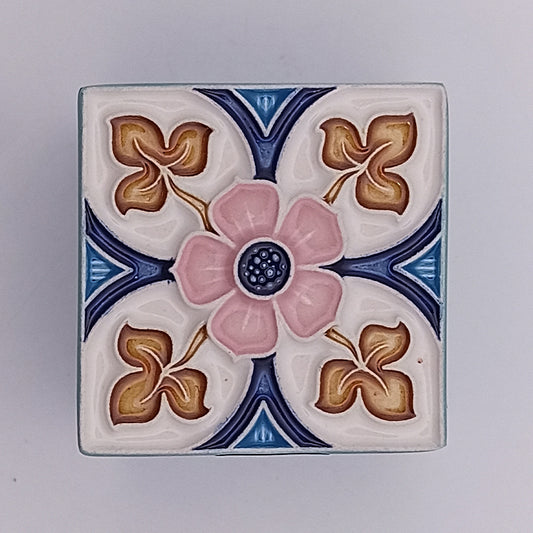 A Faro Ceramic Box by Tejo Shop with a delicately painted floral pattern. The central design is a pink flower with a dark center, surrounded by four golden-brown clover-like shapes. The background features blue and white sections with geometric accents, showcasing its expertly crafted details.