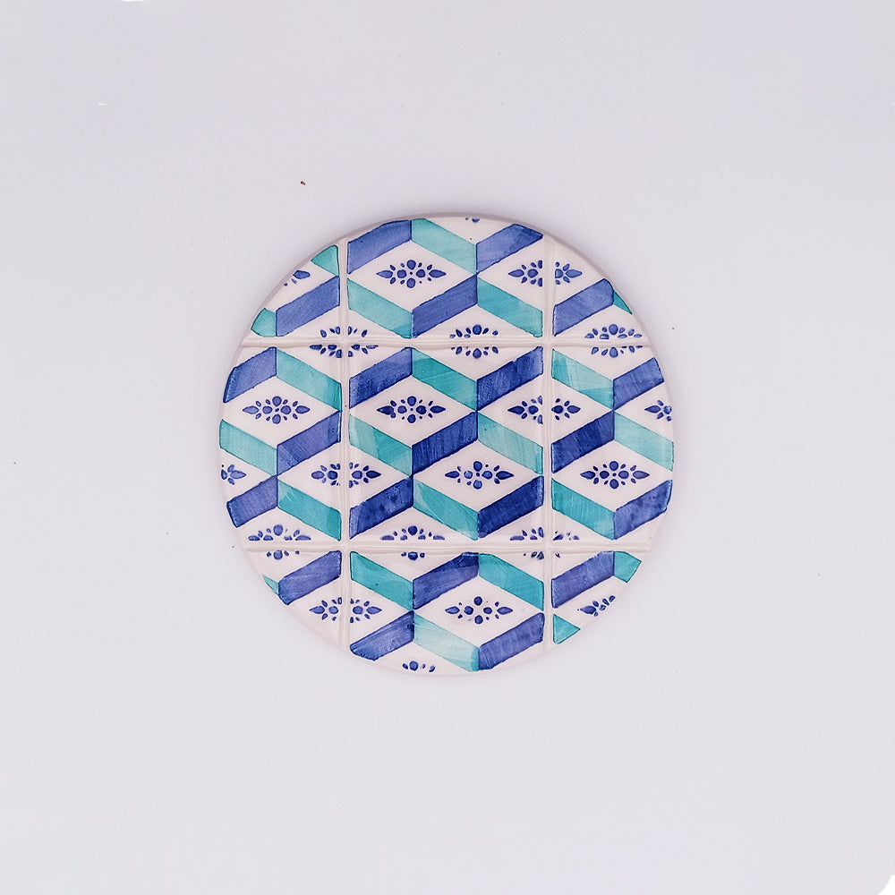 A round, hand-crafted and hand-painted coaster featuring a geometric pattern in blue, teal, and white. The design includes alternating diamond shapes and small floral motifs, reminiscent of a mosaic tile effect found in a Faro Ceramic Tile Cup Pad by Tejo Shop. The background is plain white, highlighting its intricate details.