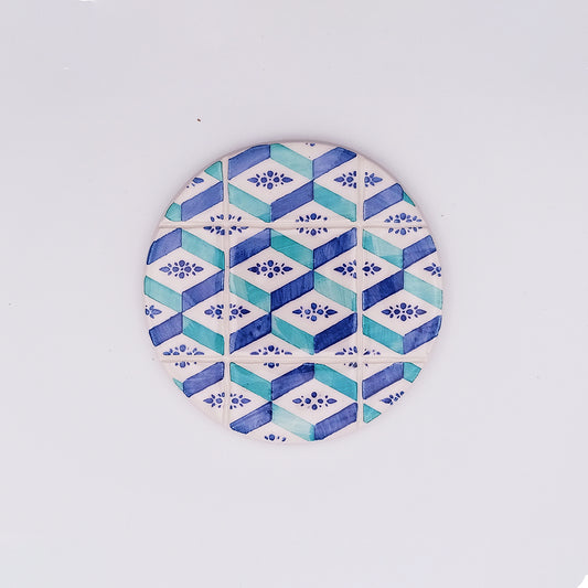 A round, hand-crafted and hand-painted coaster featuring a geometric pattern in blue, teal, and white. The design includes alternating diamond shapes and small floral motifs, reminiscent of a mosaic tile effect found in a Faro Ceramic Tile Cup Pad by Tejo Shop. The background is plain white, highlighting its intricate details.