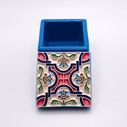 A small rectangular ceramic pot featuring artistic details in a vibrant, colorful geometric pattern on one side. The symmetrical design includes shades of pink, green, blue, and red. The top half is plain blue with an open compartment, making it an exquisite Tejo Shop Flowers Island Ceramic Box reminiscent of the Azores Islands' craftsmanship.