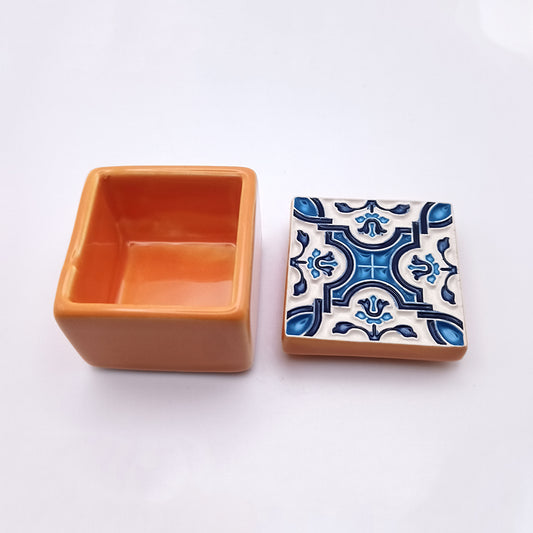 A sophisticated Funchal Ceramic Box by Tejo Shop, this small orange piece features a smooth, glossy finish and a square shape. Its lid, placed next to it, showcases intricate blue and white patterns with a central floral design. The handcrafted box stands out beautifully against the plain white background.