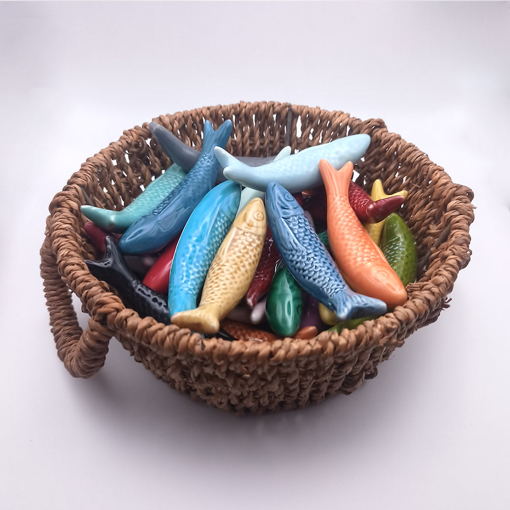 Tejo Shop's Ceramic Sardine Fridge Magnets come in a woven basket filled with vibrant ceramic fish, embodying the charm of Portuguese kitchen decor. The assortment includes colors such as blue, orange, yellow, red, green, and black. Each fish features intricately designed scales and fins that give them a lifelike appearance. Perfect for decorating a plain white fridge.