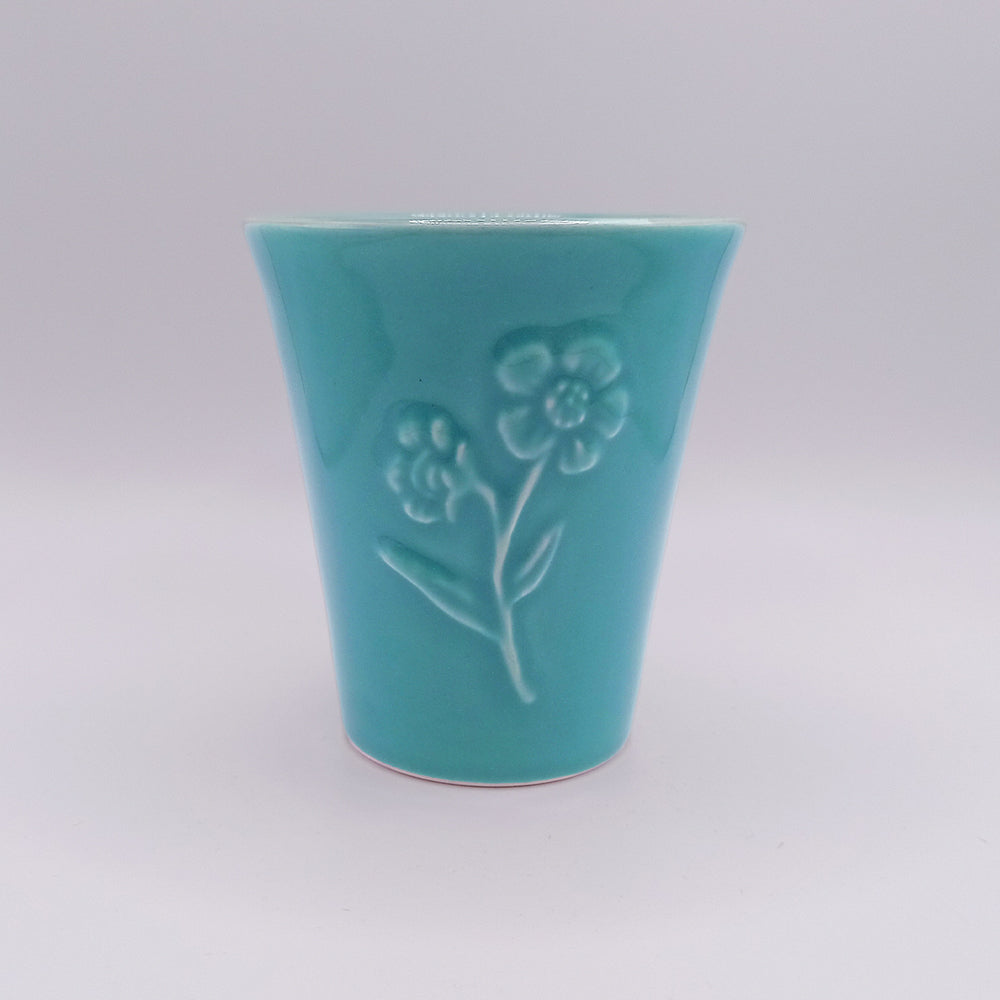 Presenting the Ceramic Mug by Tejo Shop – a unique handmade piece in turquoise with a smooth, glossy finish. This cup boasts an embossed design of two flowers with stems and leaves on its front, capturing the charm of Portuguese art. The plain white background elegantly highlights the intricate details.