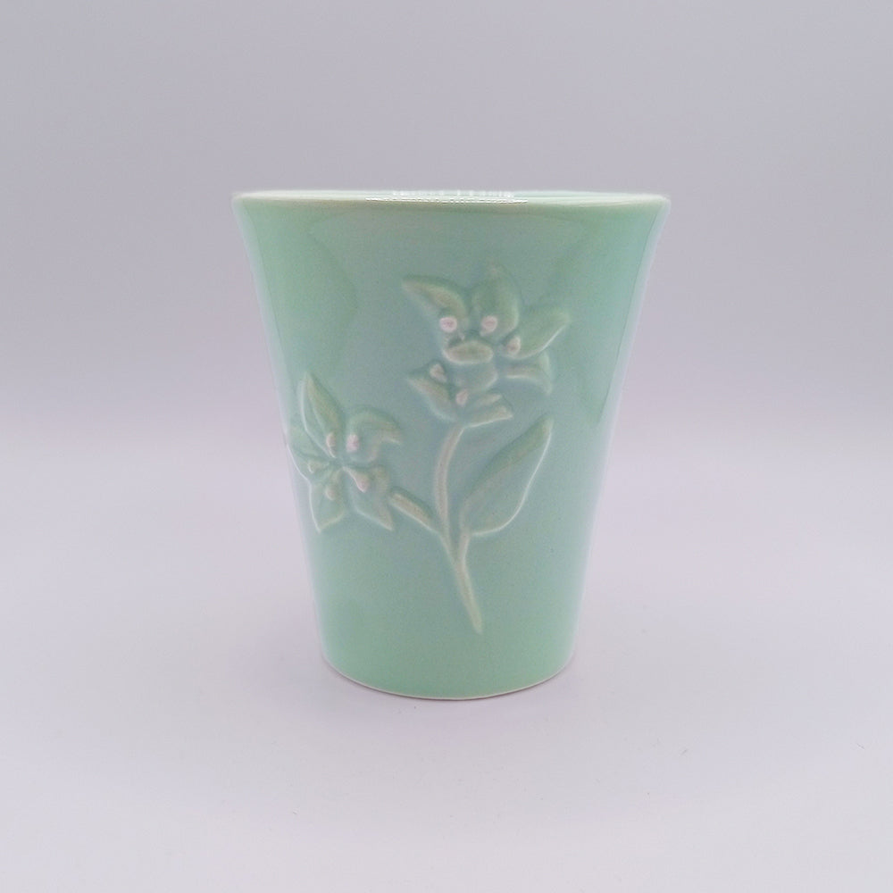 A light green ceramic mug by Tejo Shop with a subtle embossed floral design featuring two flowers and leaves on the front. The mug, inspired by Portuguese art, has a slightly flared top and a smooth, glossy finish. The background is plain and white, highlighting the mug's intricate craftsmanship.