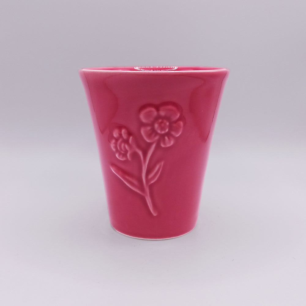 The Ceramic Mug from Tejo Shop is a pink ceramic cup with a glossy finish, adorned with an embossed flower and leaf pattern on its side. Influenced by Portuguese art, this unique mug has a slightly flared shape and a smooth surface, displayed against a light gray background.