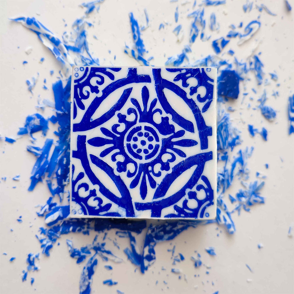 The Amêndoa Royale Tile Soap Bar by Lisbão, featuring a handcrafted square design with intricate blue floral and geometric patterns, rests on a white surface surrounded by scattered blue and white fragments.