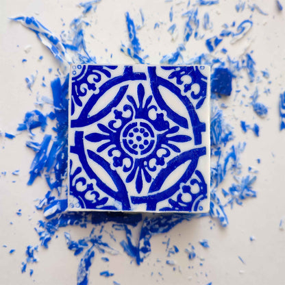 The Amêndoa Royale Tile Soap Bar by Lisbão, featuring a handcrafted square design with intricate blue floral and geometric patterns, rests on a white surface surrounded by scattered blue and white fragments.