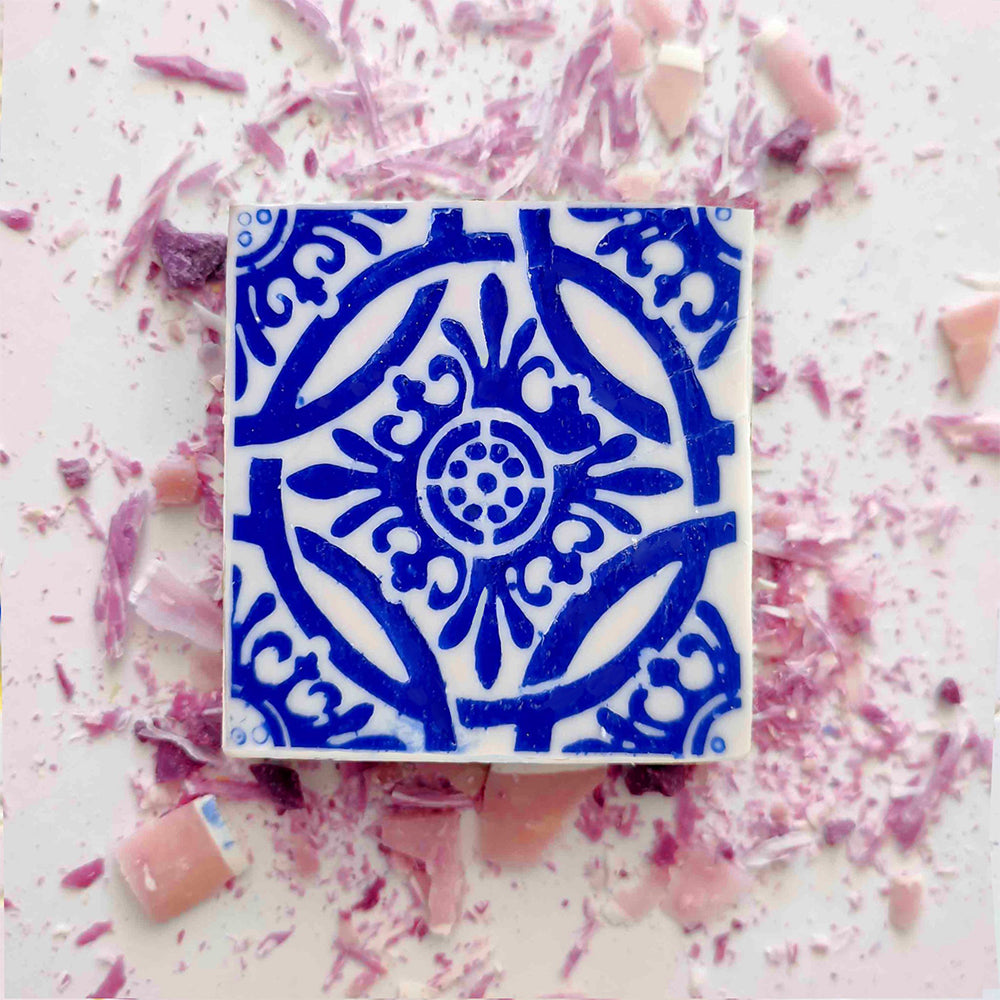 A square Lisbão Rosa de Azulejo handmade Portuguese tile soap, featuring an intricate blue pattern, rests on a white surface amid scattered pink and purple candy shavings and small broken pieces.