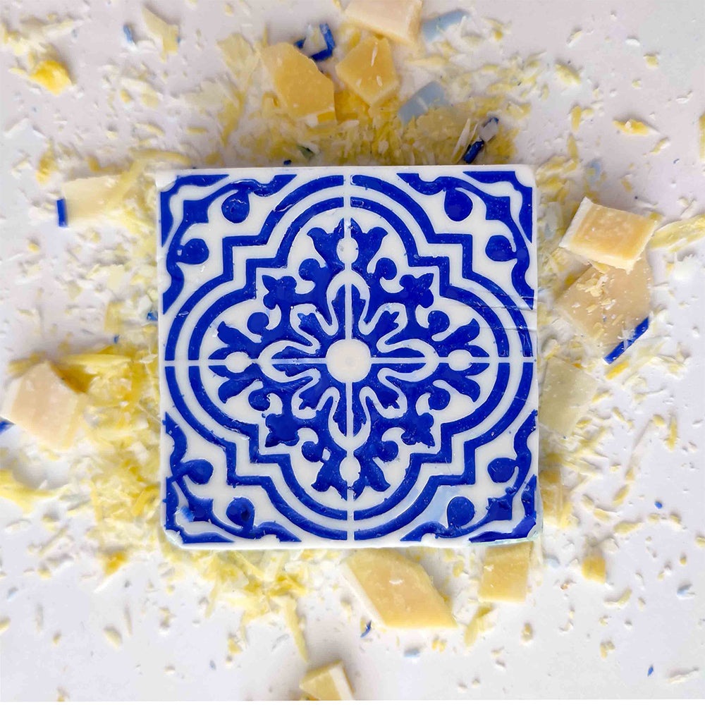 The Lisbão Pastel de Azulejo vegan soap bar has a blue and white geometric tile pattern. It sits on a white surface, surrounded by yellow and white soap shavings.