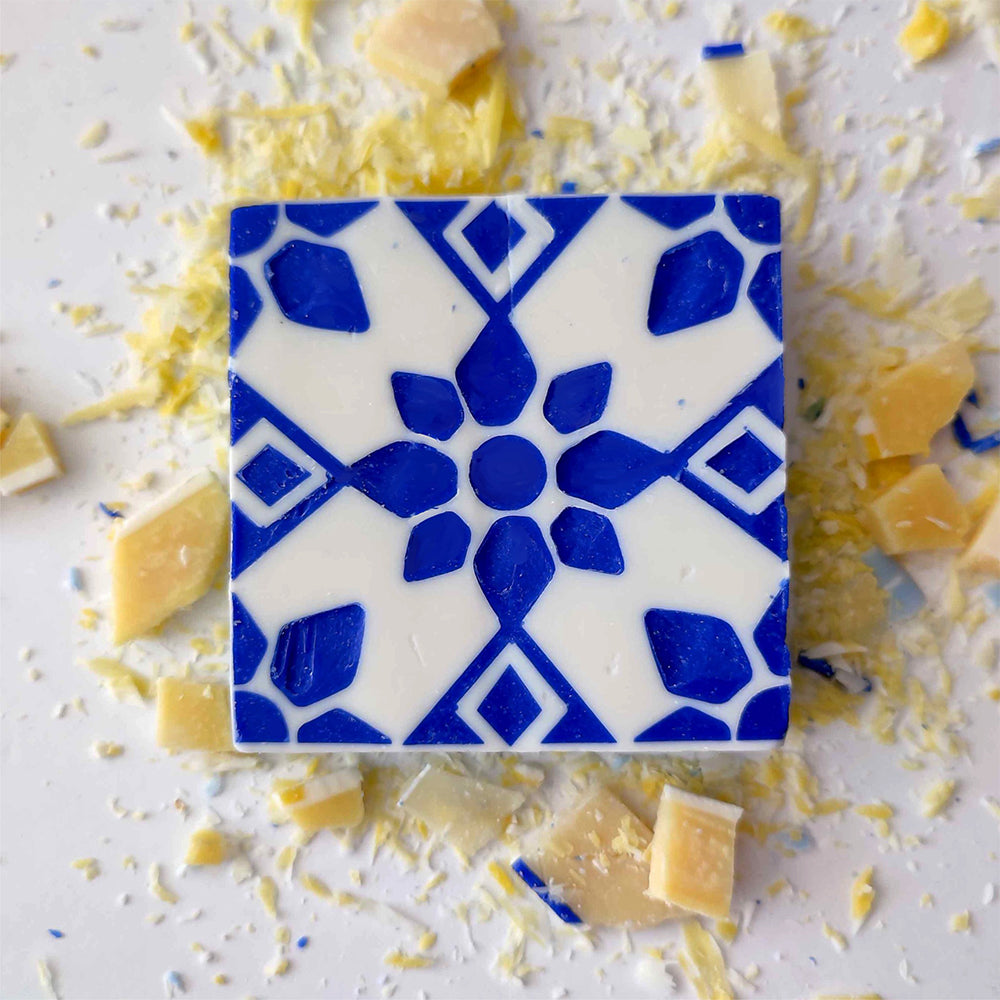 Pastel de Azulejo by Lisbão features a square soap with a blue geometric floral pattern on white, accented by yellow and beige shards—handmade and inspired by classic Portuguese azulejo tile designs for a unique touch.