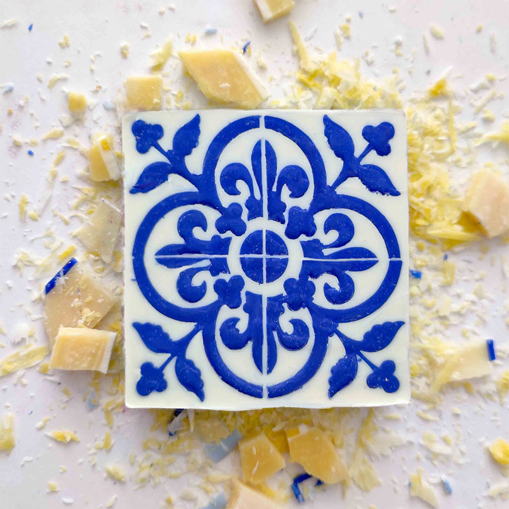 The Lisbão Pastel de Azulejo is a square white Portuguese tile soap with a blue ornate floral and geometric design, displayed among grated cheese and cheese chunks on a white surface.