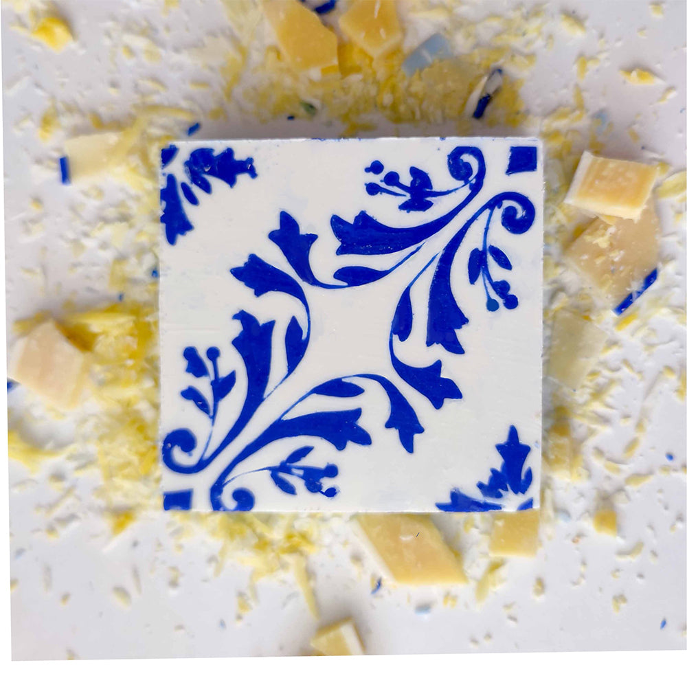 The Pastel de Azulejo soap by Lisbão features a white square shape with a blue floral pattern, displayed on a white surface and surrounded by scattered white and yellow chocolate shavings and small chunks.