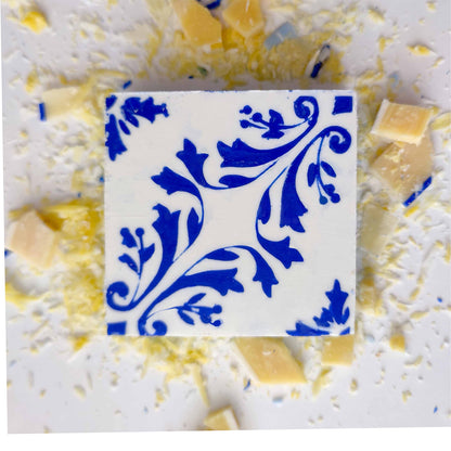 The Pastel de Azulejo soap by Lisbão features a white square shape with a blue floral pattern, displayed on a white surface and surrounded by scattered white and yellow chocolate shavings and small chunks.