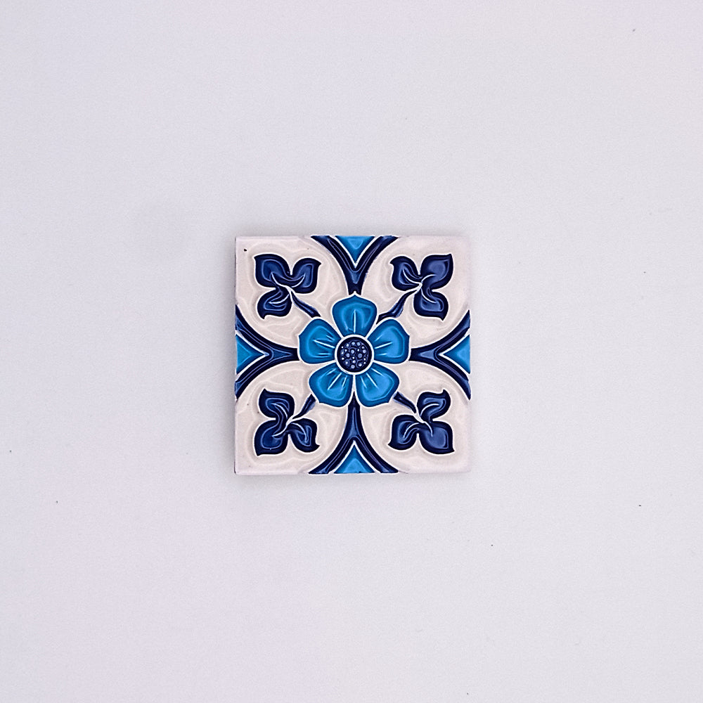 A decorative Tejo Shop Royal Blue Small Ceramic Tile featuring a symmetrical floral design in shades of blue and white, centered with a raised blue button-like detail, against a plain light background.