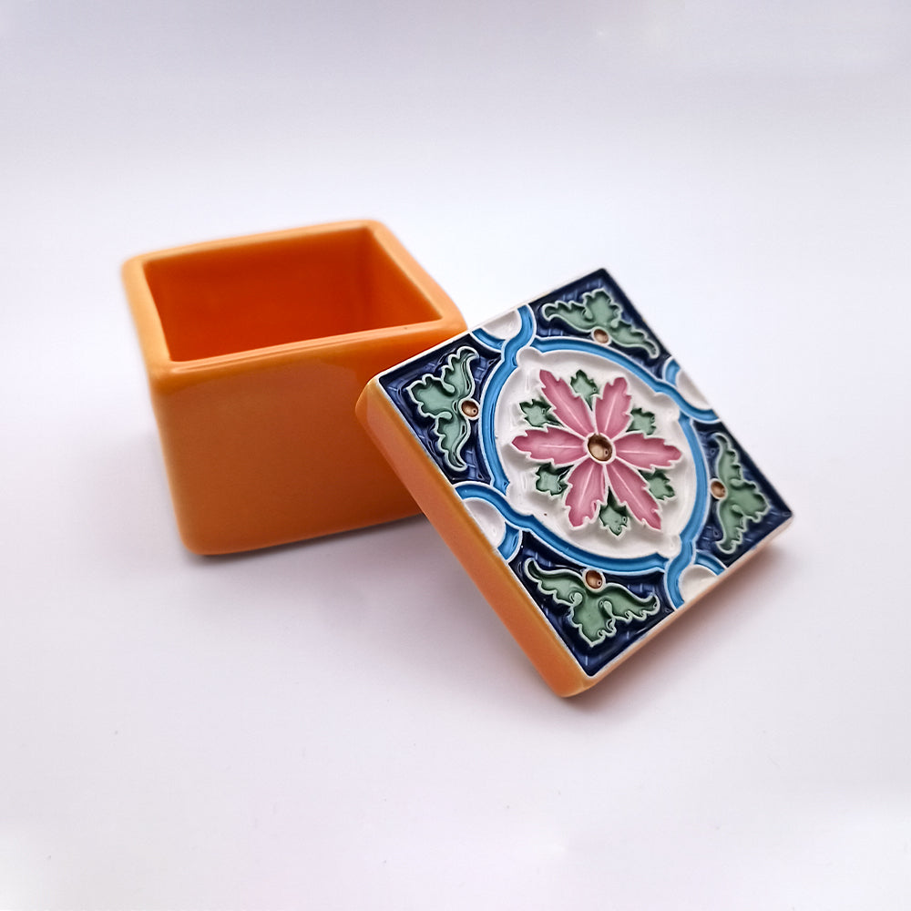 A small orange square box with an ornate lid slightly ajar. The Horta Island Ceramic Box by Tejo Shop features a colorful, intricate floral pattern with pink, green, and blue designs on a white background. The exquisite ceramic box has a smooth, polished finish and is set against a solid white backdrop.