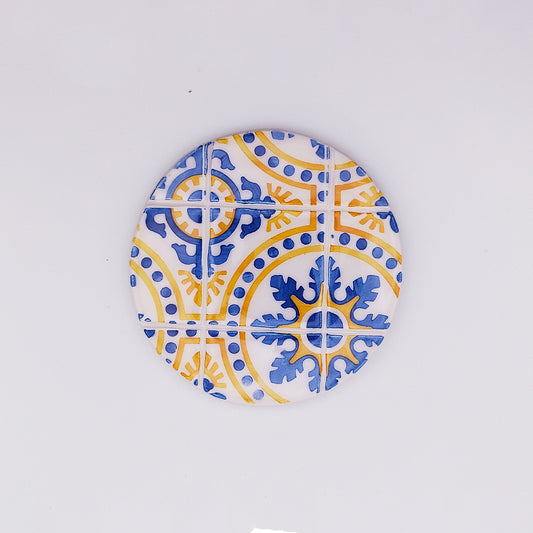 The **Island of Madeira Ceramic Tile Cup Pad** by **Tejo Shop** features a mosaic design with blue and yellow motifs on a white background. The pattern includes intricate floral and geometric shapes, resembling traditional tile artwork, adding artisanal charm to any setting.