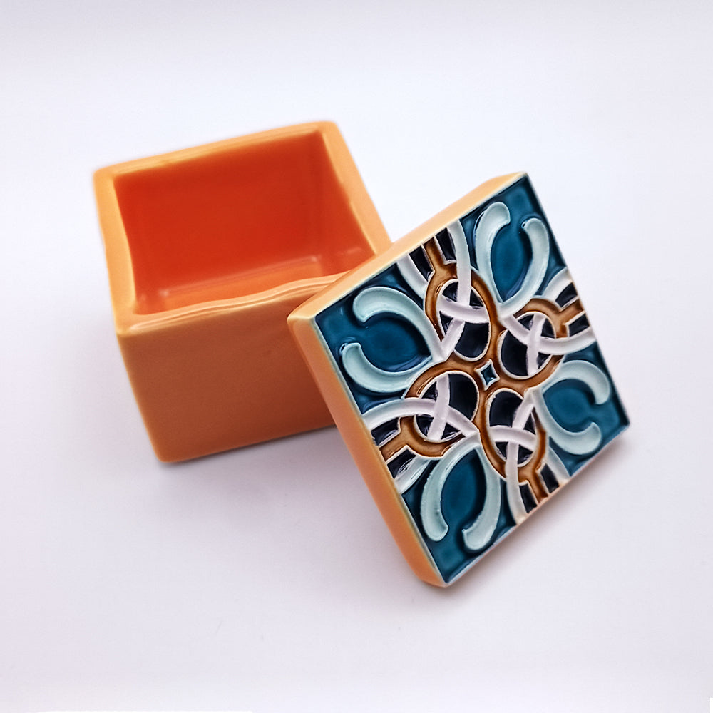 A small, square, orange ceramic marvel with an open lid placed beside it. The lid features an intricate tile design with interlocking patterns in blue, white, and brown colors. The luxury interior of the Leiria Ceramic Box by Tejo Shop is also orange. The background is solid white.