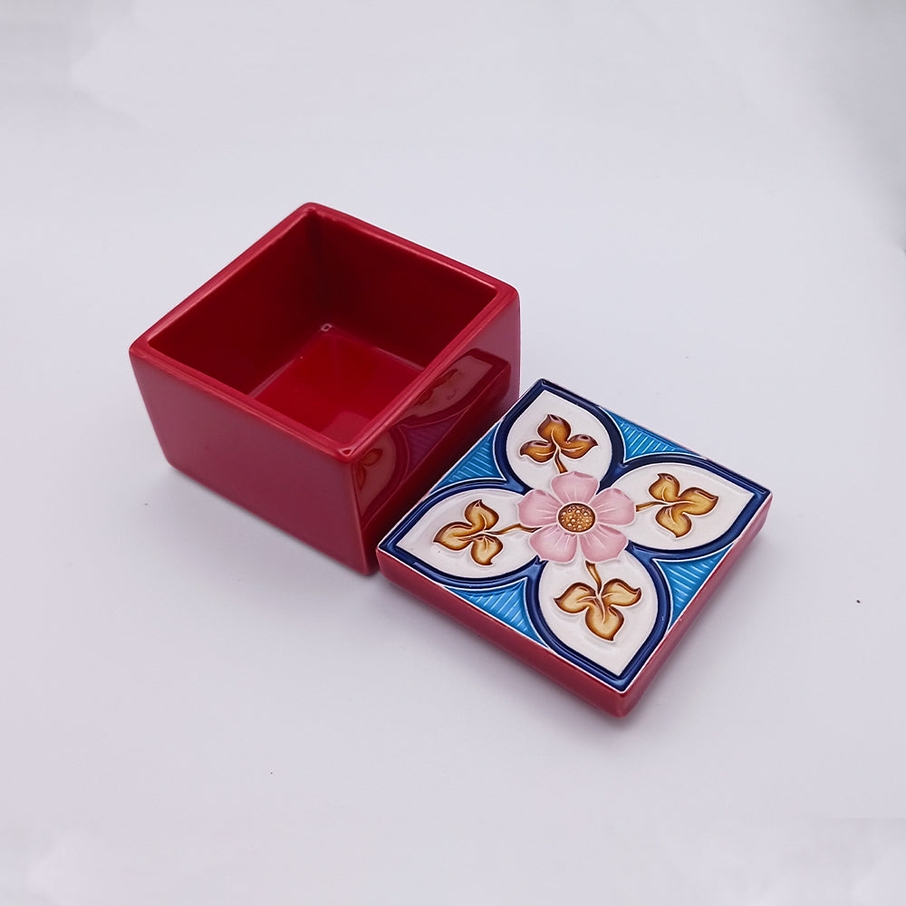 A small, red, square ceramic box with its lid removed. The lid is decorated with a floral design featuring a central pink flower surrounded by blue and gold petal-like patterns. The interior of the box is plain red. This is the Liberdade Ceramic Box from Tejo Shop.