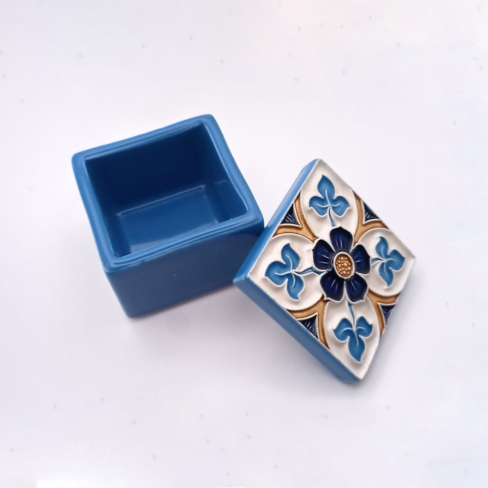 A small, square, blue ceramic box with a detached lid. This exquisite Madeira Island Ceramic Box by Tejo Shop features an intricate hand-painted design with a blue flower pattern, blue leaves, and gold accents against a white background. The background surface is white with a subtle texture.