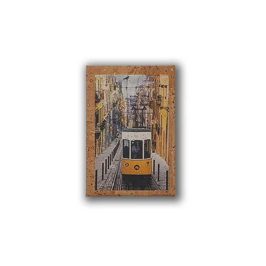 The Bica - Cork Fridge Magnet by New Jamp captures a yellow tram climbing a steep, narrow Lisbon street with colorful buildings and overhead wires, seen through a rain-speckled window—an iconic city souvenir.