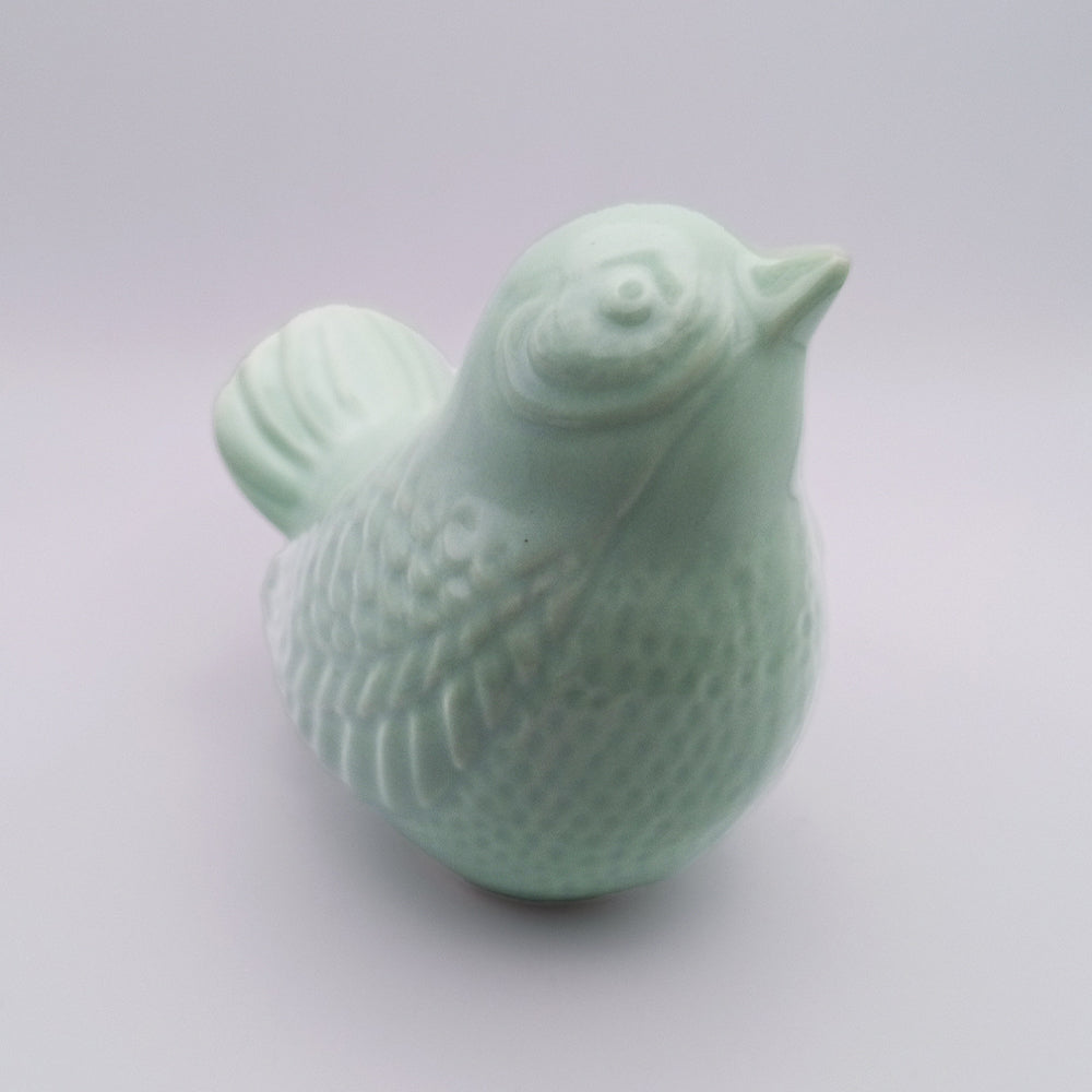 The "Ceramic Bird looking up Figurine" by Tejo Shop is a handmade, light green ceramic bird figurine featuring detailed feathers and a smooth, glossy finish. Its pointed beak and prominently crafted eyes stand out against the plain, out-of-focus background, highlighting the intricate design of this exquisite piece of home decor.