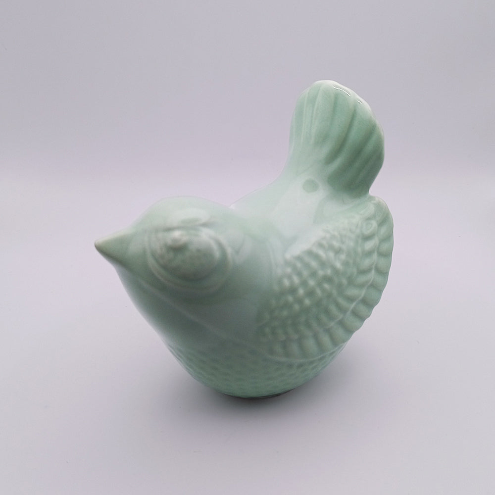 The Ceramic Bird Figurine by Tejo Shop is a small, smooth porcelain ceramic featuring a light mint-green color. This handmade bird showcases detailed feather engravings, a prominent tail, and rounded eyes, all set against a plain white background. It's perfect for adding charm to your home decor.