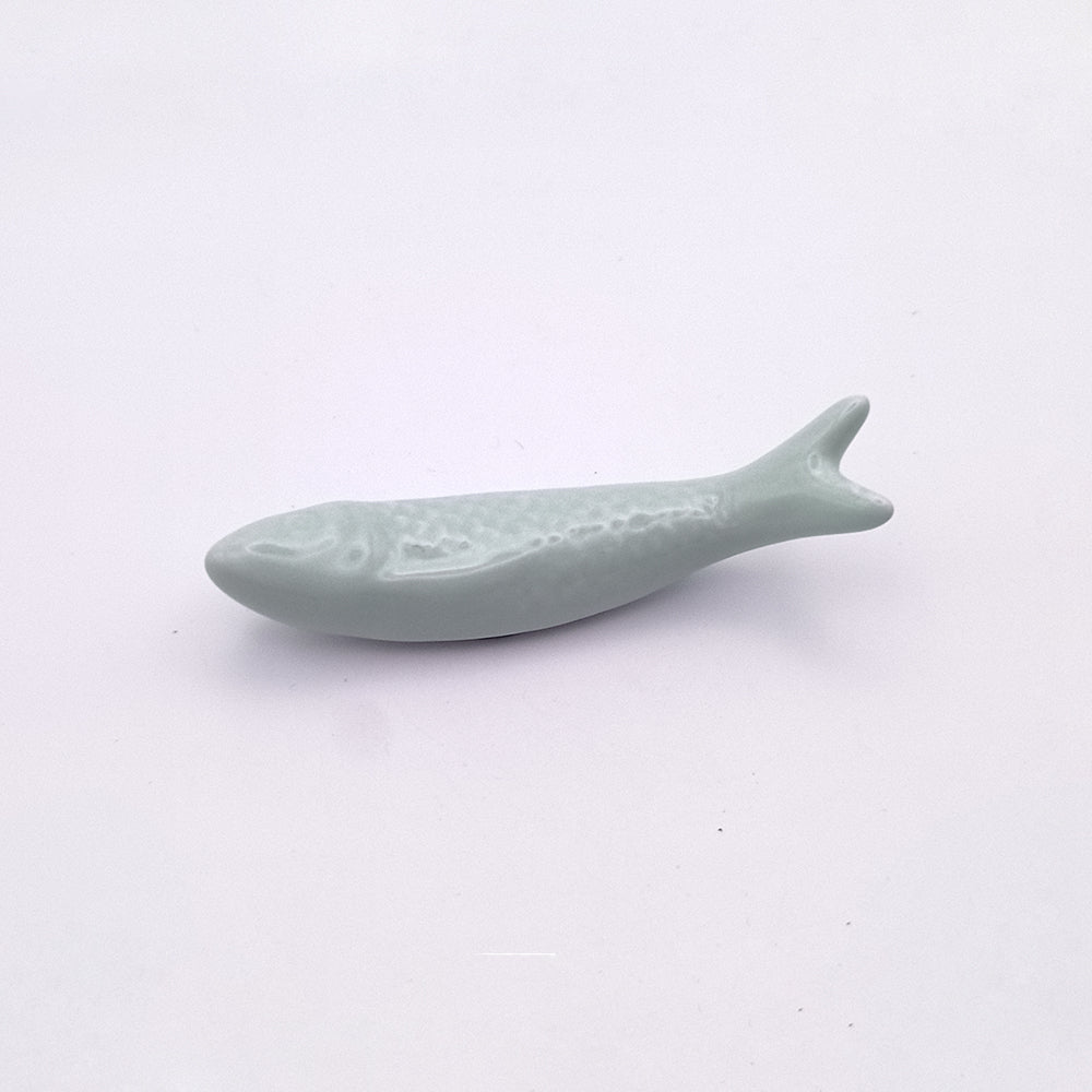 A small, light blue Ceramic Sardine Fridge Magnet by Tejo Shop features a smooth texture and slightly raised scales. Viewed against a plain white background, this Portuguese icon is positioned horizontally with its head on the left and tail on the right, making it perfect for adding charm to your kitchen decor.