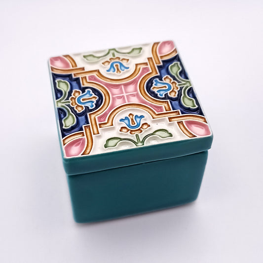 A small, square ceramic box with a teal base and a lid featuring artistic details of floral and geometric designs in shades of green, blue, pink, and gold. This Oporto Ceramic Box by Tejo Shop adds home sophistication, set against a plain white background.