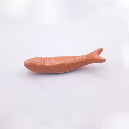 A smooth, glossy Ceramic Sardine Fridge Magnet by Tejo Shop lies on a white background. This small, salmon-colored fish-shaped object resembles a popular Portuguese icon and features textured scales and fins, making it an eye-catching piece of kitchen decor.