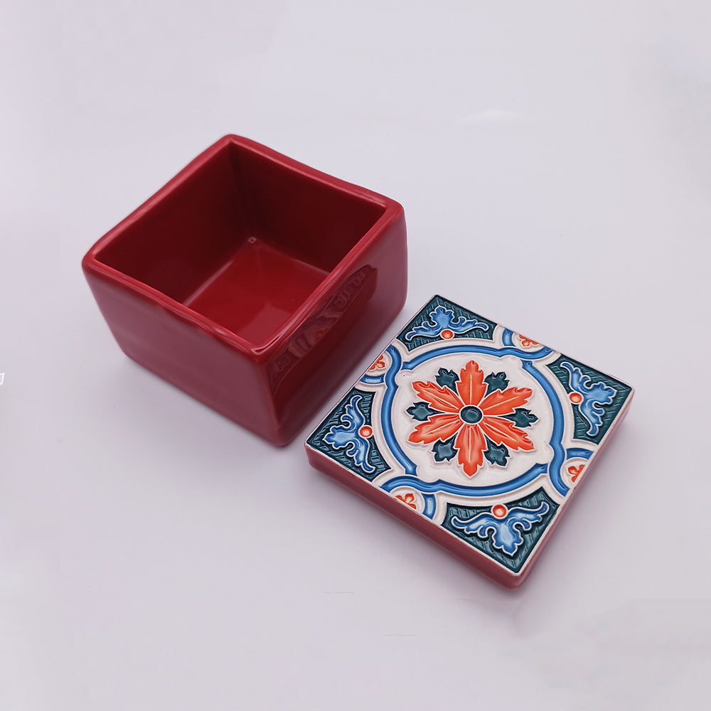 A sophisticated home decor piece, the Portimão Ceramic Box by Tejo Shop is a square red ceramic box with an open lid lying on a white surface. The lid boasts an intricate floral pattern in red, orange, blue, and white, resembling traditional tile design. The interior of the handcrafted box is solid red.