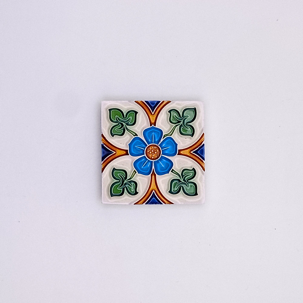 A decorative ceramic tile featuring a colorful floral design with blue, green, orange, and white patterns centered around a blue dot on a gray background, reminiscent of Tejo Shop's Spanish Style Tiles.