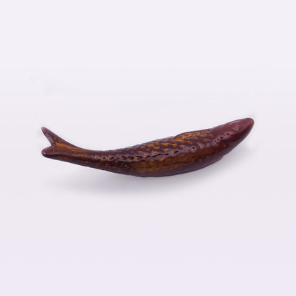 A close-up of a brown, elongated fish-shaped object with textured scales, photographed against a plain white background. This handcrafted Portuguese Ceramic Sardines by Tejo Shop appears as a detailed, artistic representation of a fish with a pointed head and a slightly curved body and tail.