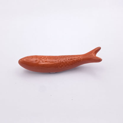 A small, smooth, brown ceramic object shaped like a fish with textured details on its body and fins rests flat on a plain white surface, perfectly capturing the essence of a Portuguese icon. This charming piece from Tejo Shop doubles as their Ceramic Sardine Fridge Magnet, making it ideal for stylish kitchen decor.