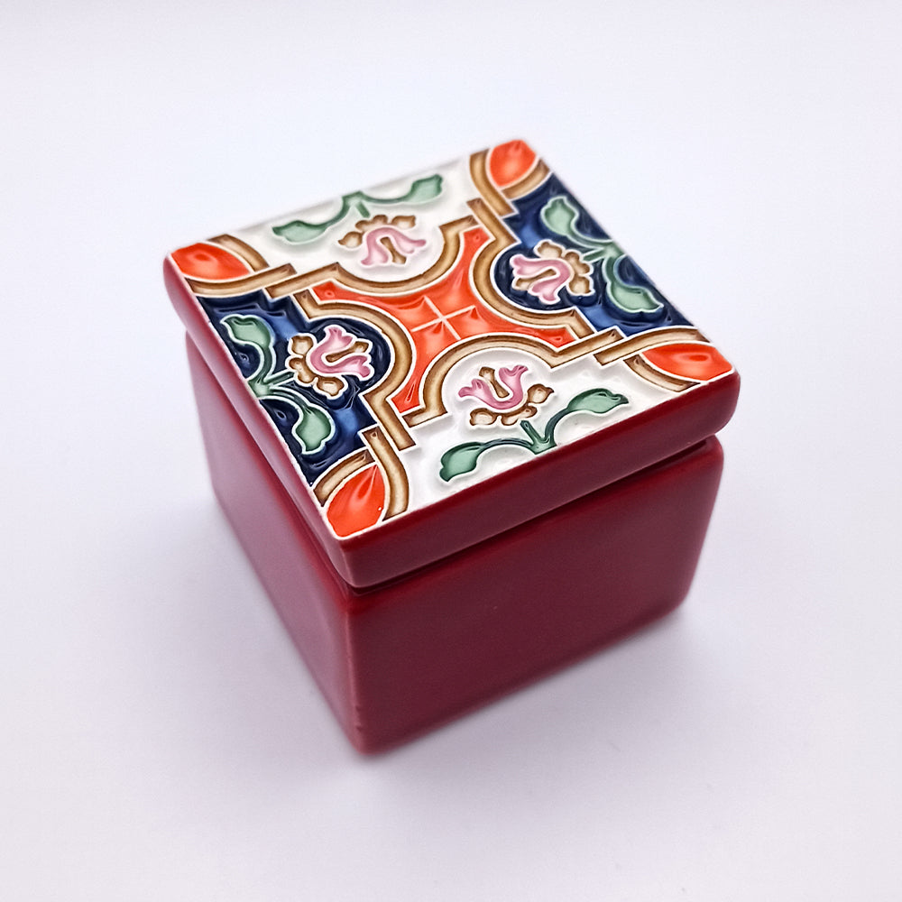 A small, square, red ceramic box with a detachable lid. The Rossio Ceramic Box by Tejo Shop features a colorful, intricate pattern with floral and geometric designs in shades of green, blue, red and orange. This exquisite piece of home decor is set against a plain white background.